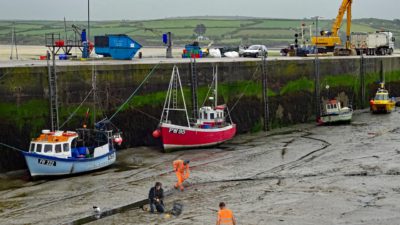 En hamn utan vatten. Längs med hamnkanten ligger det flera båtar på bottnen. På bottnet står flera personer i reflexkläder och gräver i gyttjan. Personerna ser lite konstiga ut i bilden, som att de är inklippta.