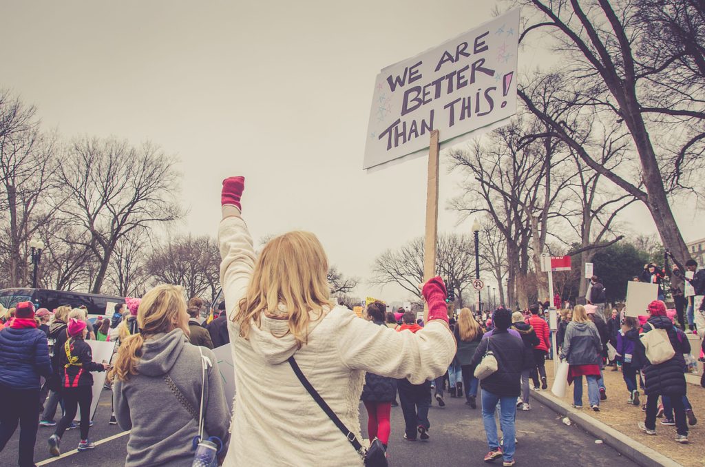 Bilden visar en manifestation för kvinnor. På bilden syns en person som håller i en skylt och knyter näven i luften. Runt om henne går personer iklädda rosa mössor. Mössorna är en symbol för tjejer och kvinnors rättigheter.